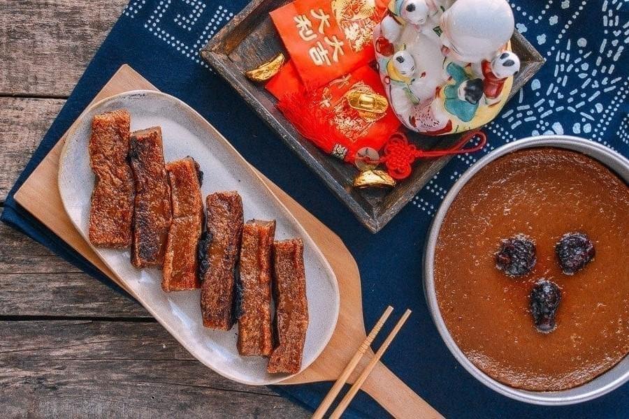  Gemini said The image displays a festive spread featuring Nian Gao, a sticky lunar new year cake, presented both in a whole round tin and as pan-fried slices on a long platter. Surrounding the food are traditional Chinese decorations, including red envelopes (hongbao), gold ingots, and a porcelain figurine, all set against a rustic wooden background.