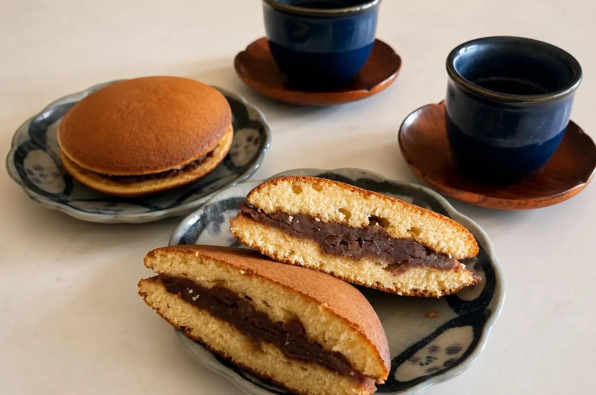 Two plates of dorayaki (Japanese red bean pancakes) served on a white tabletop alongside two small, dark blue cups of tea on wooden saucers. In the foreground, one dorayaki is sliced in half, revealing the thick sweet red bean paste filling between the fluffy, golden-brown pancakes.