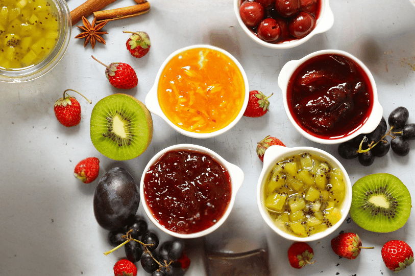 Five small bowls of jewel-toned fruit preserves—kiwi, cherry, marmalade, plum, and berry—sit surrounded by fresh fruits and warm spices, illustrating the vibrant possibilities of glazing in fruit tart preparation.