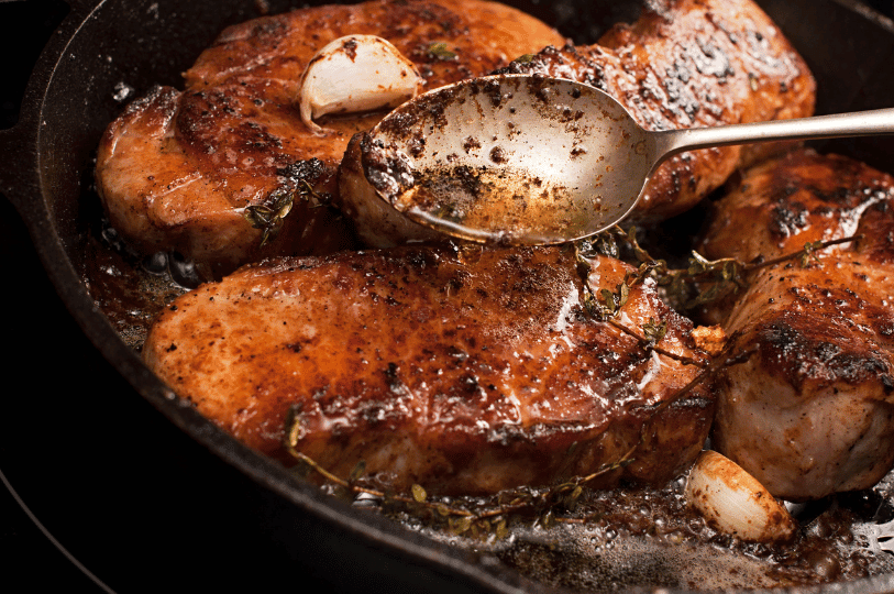 Several pork chops being butter-basted in a cast-iron pan with garlic cloves and thyme to enhance the flavor of the seared crust.