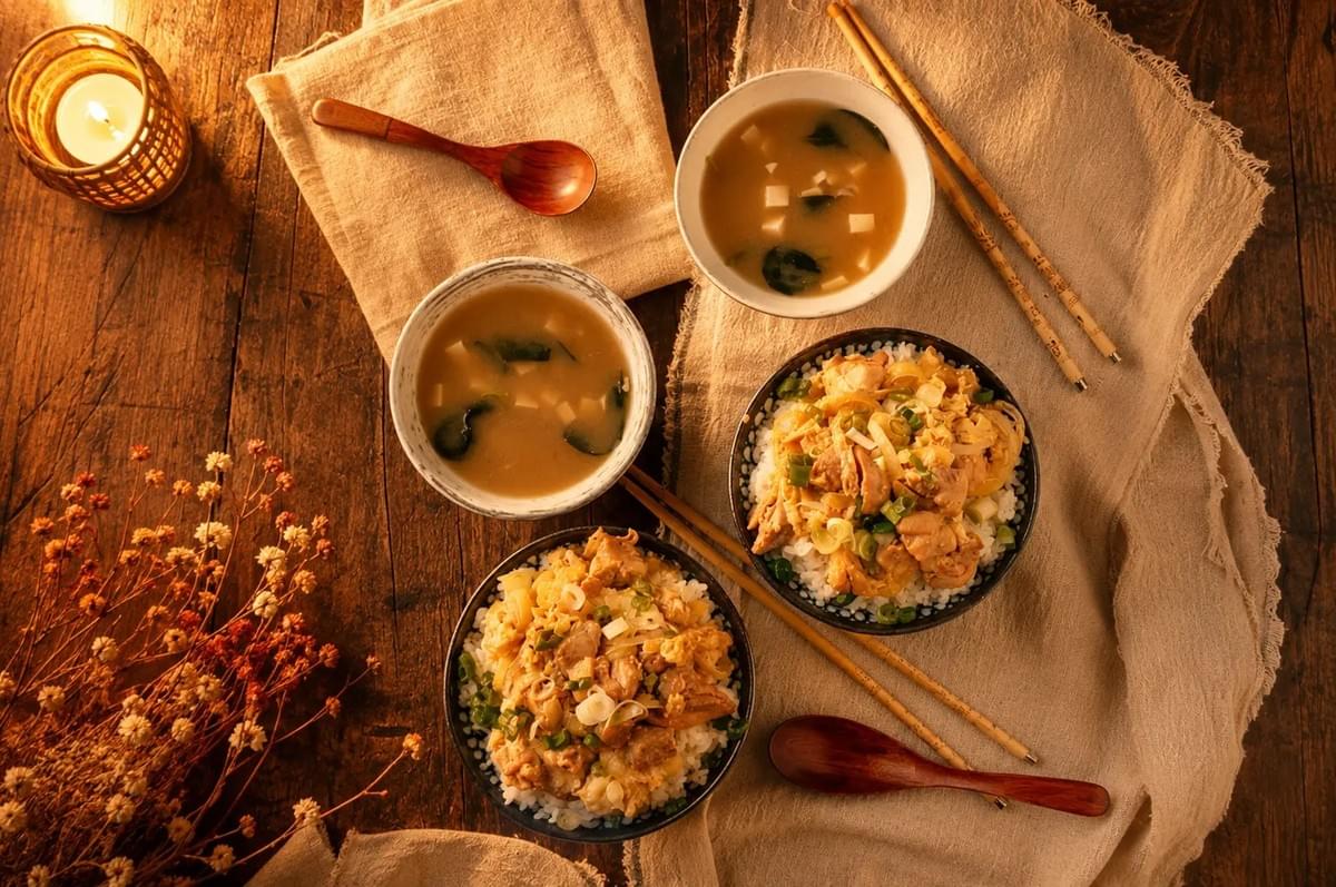 A top-down shot of a Japanese dinner setting for two on a wooden table. It features two bowls of Oyakodon (chicken and egg rice), two bowls of miso soup, wooden spoons, chopsticks, linen napkins, and a lit candle.