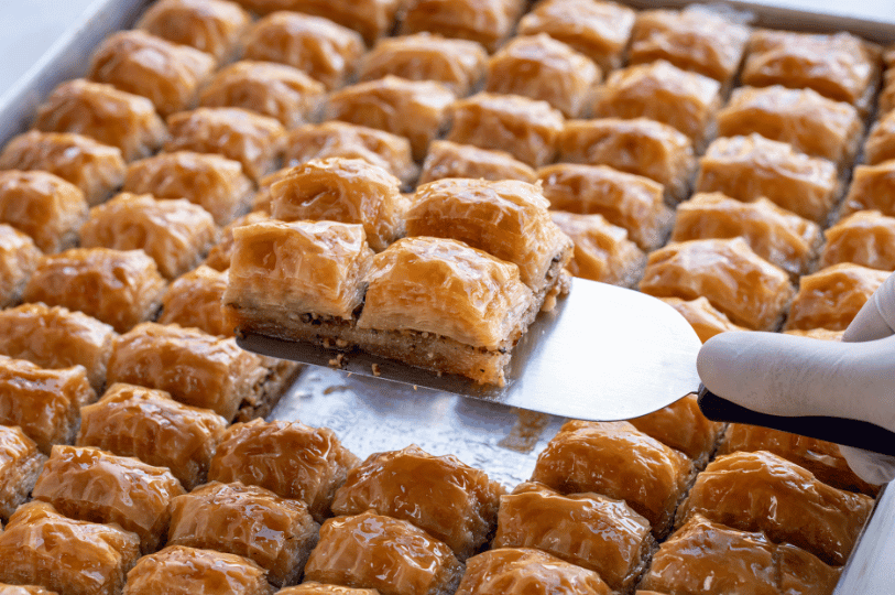 A metal tray filled with rows of golden-brown baklava, with a gloved hand using a spatula to lift out a crisp, honey-soaked square to show the dense nut filling.
