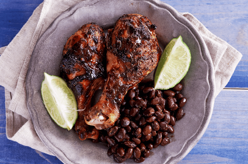 Two glazed jerk chicken drumsticks served on a rustic pewter plate alongside a portion of seasoned black beans and fresh lime wedges, set against a blue wooden background.