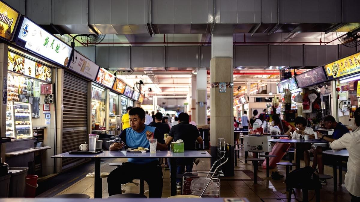 This image shows the interior of a bustling Singaporean hawker centre, where patrons are seated at tables amongst rows of various food stalls. In the foreground, a man sits alone at a table, focused on his phone while enjoying a meal and a cold drink.