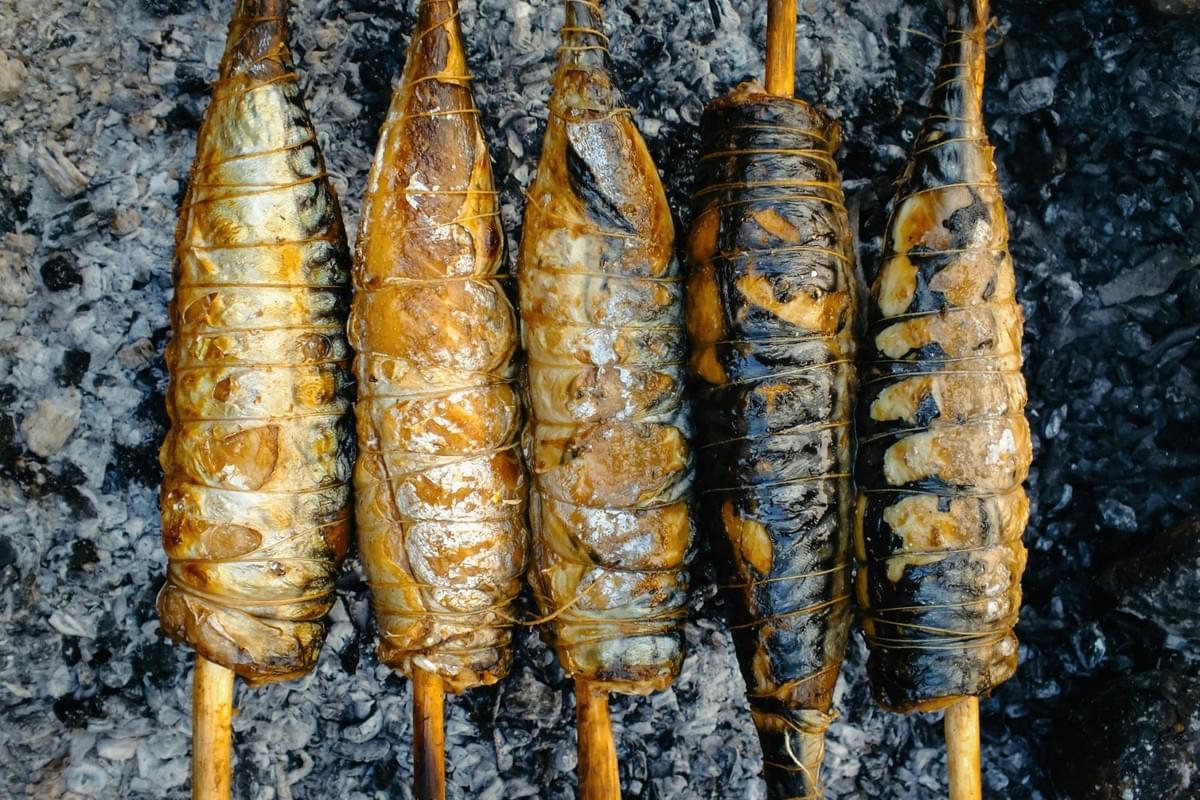 This overhead, close-up photograph captures five whole fish skewered lengthwise on wooden sticks and being roasted over hot charcoal embers.