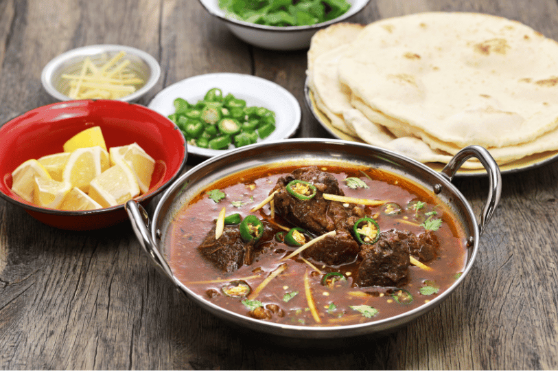 A traditional Pakistani Nihari served in a metal bowl, surrounded by essential garnishes including lemon wedges, sliced green chilies, and fresh ginger strips, with a stack of warm naan in the background.