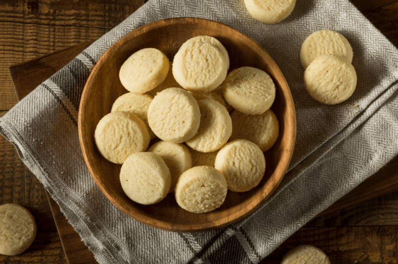 An overhead shot of a wooden bowl filled with round shortbread cookies, resting on a grey linen napkin and a rustic wooden table with a few cookies scattered nearby.