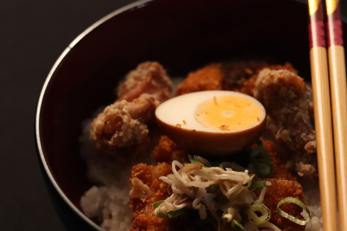 A high-angle, close-up shot shows a Japanese ramen or rice bowl filled with rice, crispy fried meat, bean sprouts, chopped scallions, and a halved soft-boiled ramen egg. A pair of wooden chopsticks rests along the right edge of the dark bowl on a dark background.