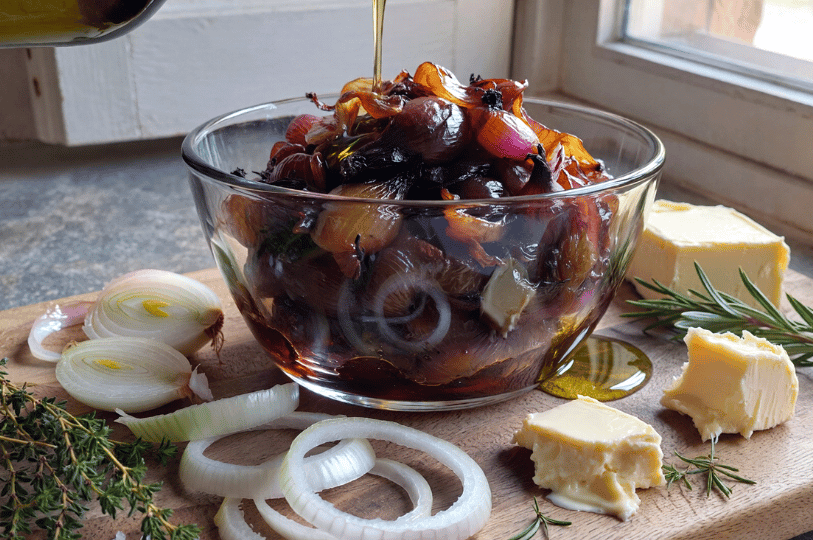 A glass bowl filled with warm caramelized onions sits on a wooden counter next to blocks of butter, fresh rosemary, thyme, and raw onion slices, with olive oil being drizzled over the top.