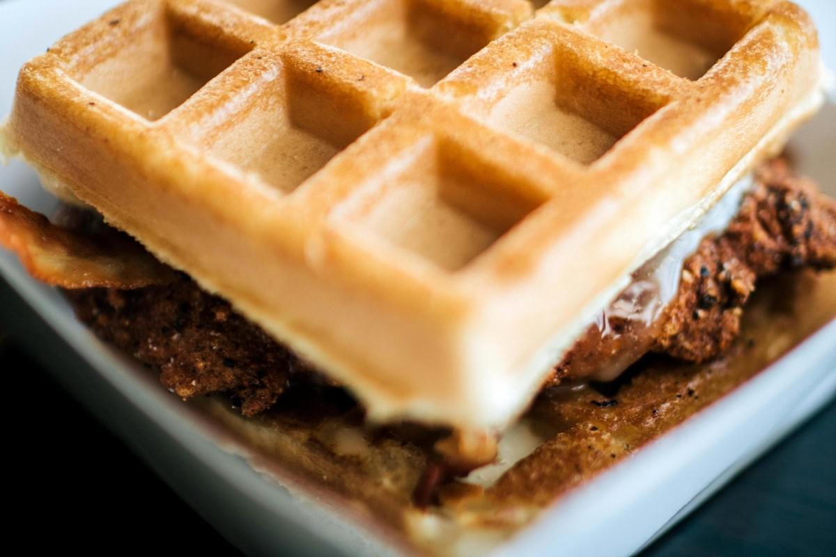 A close-up view of a chicken and waffle sandwich featuring a crispy, golden-brown fried chicken piece layered with bacon between two waffles. Glazed syrup or sauce drips lightly along the edge, highlighting the savory and sweet dish.