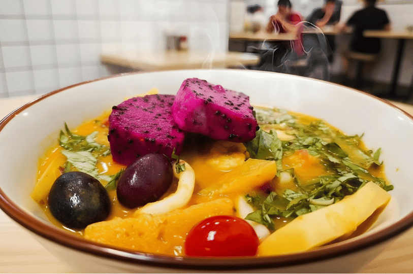 A detailed shot of a bowl of fruit-infused curry, showcasing vibrant purple dragon fruit, grapes, and cherry tomatoes swimming in a thick, orange broth. Steam rises from the hot dish, which is garnished with fresh green herbs.