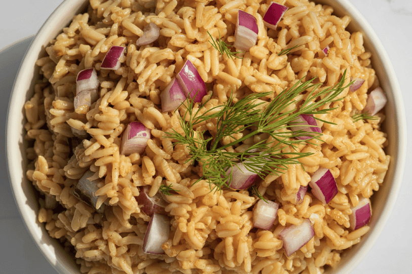 A top-down view of golden rice pilaf served in a plain white bowl, freshly garnished with diced red onions and a sprig of dill.
