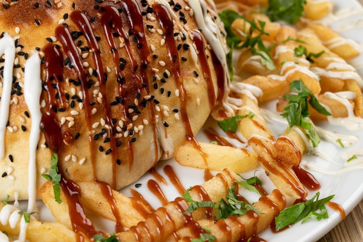 A golden-brown dome-shaped pie or stuffed flatbread sits on a plate, sprinkled with white and black sesame seeds and drizzled with white and brown sauces. Golden french fries are scattered around it, also drizzled with sauces and garnished with chopped fresh herbs.