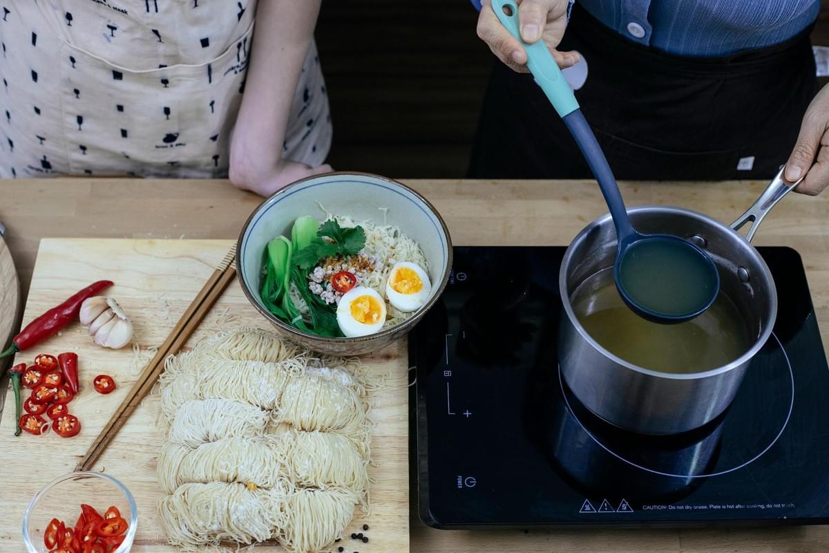 An overhead view of the image shows a person using a blue ladle to scoop clear broth from a stainless steel pot on an induction cooktop. Next to the cooktop, a wooden board holds a prepared bowl of ramen noodles with soft-boiled eggs, bok choy, minced meat, raw noodle nests, and sliced red chilies.