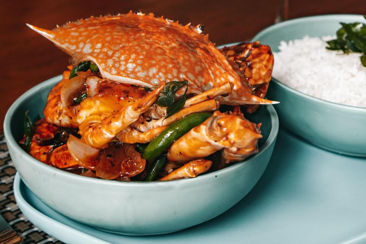 A cooked whole crab tossed in a rich, savory sauce with onions and herbs is served in a light blue bowl. Next to it sits a separate bowl filled with fluffy white rice topped with green garnishes.