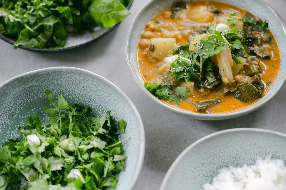 A bowl of hearty vegetable curry topped with fresh cilantro and sesame seeds sits alongside sides of white rice and chopped greens. The vibrant meal is presented in textured ceramic bowls on a clean, minimalist surface.