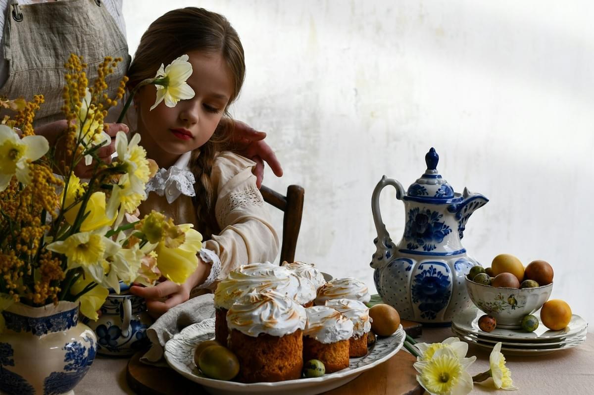 A young girl sits at a table, staring at a plate of colorful cupcakes, evoking feelings of comfort and nostalgia.