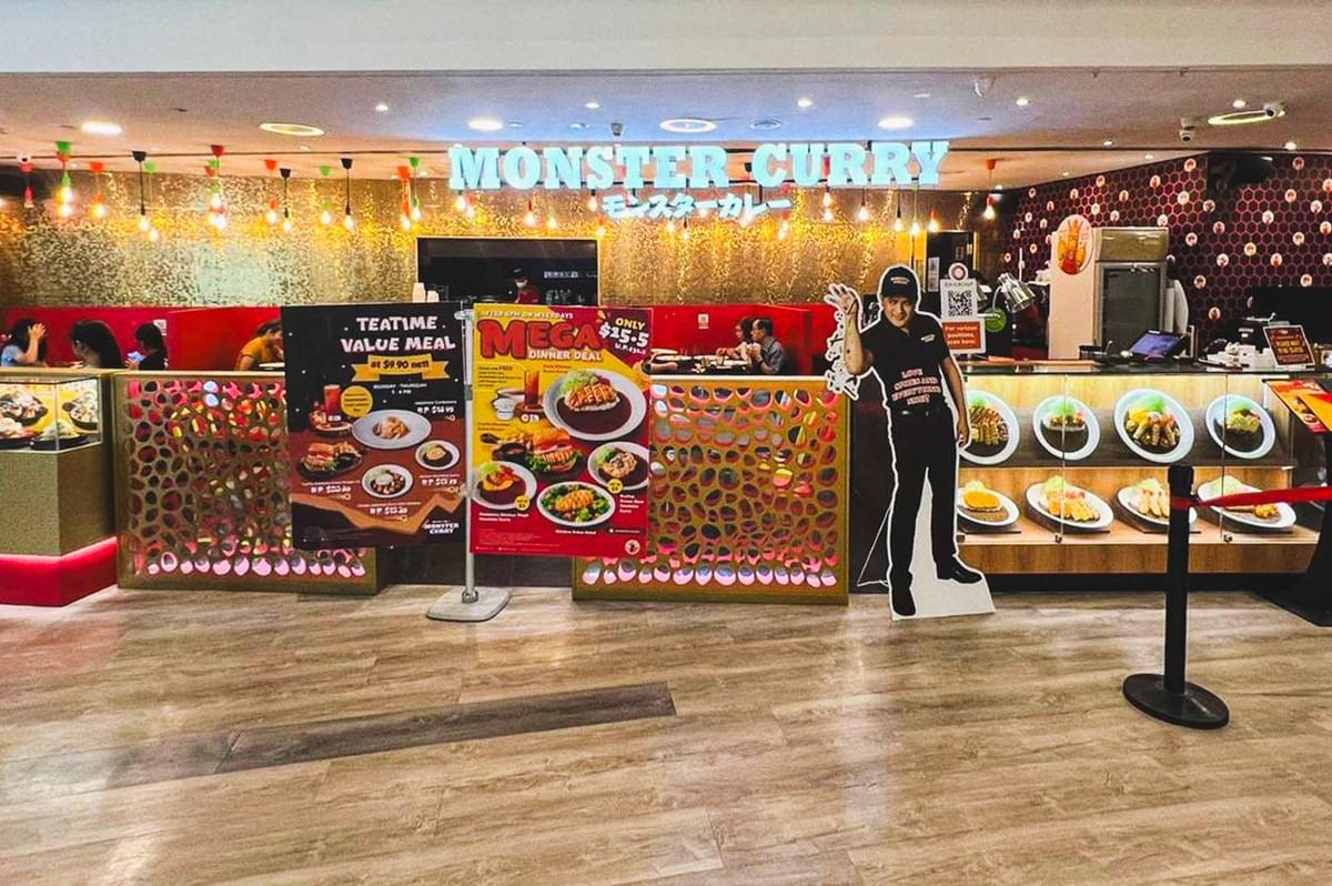Exterior of Monster Curry restaurant featuring illuminated signage, promotional posters, dish displays, and a vibrant interior with red seating and decorative lighting.