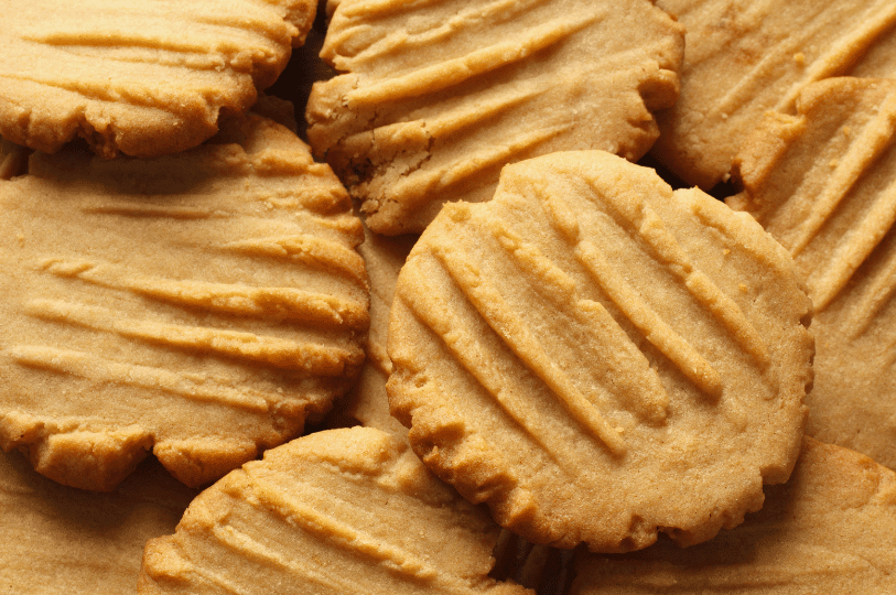 A close-up shot of several golden-brown peanut butter cookies stacked on a dark surface, showing the classic crisscross fork pattern and a crumbly, soft texture.