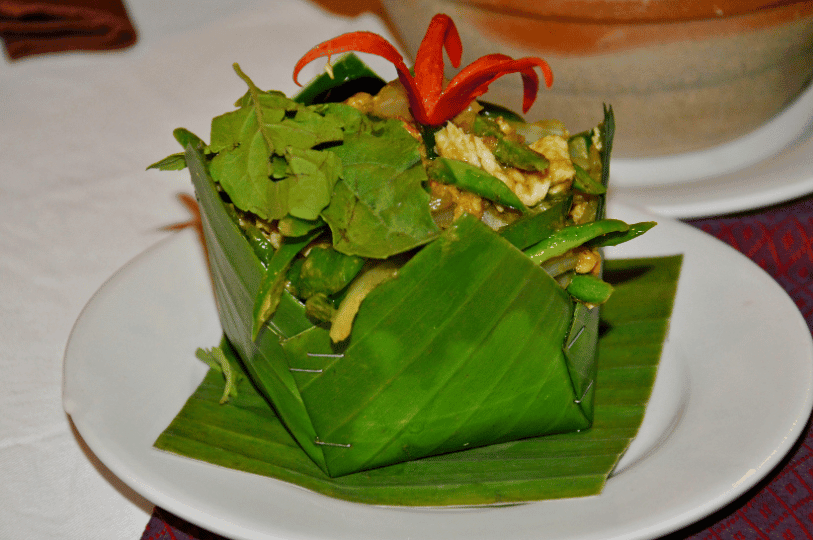 An overhead close-up of vibrant yellow steamed fish curry in a dark bowl, perfectly finished with a swirl of white coconut cream, shredded green lime leaves, and thin slices of red chili.