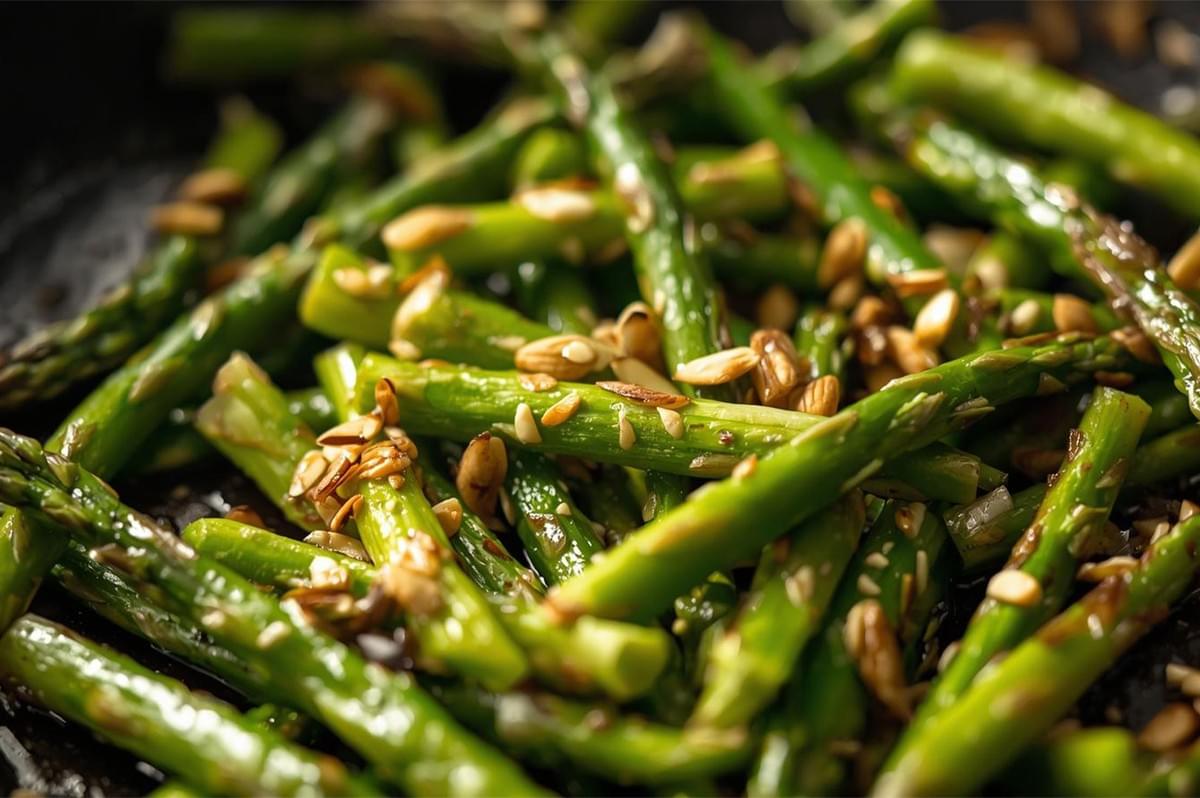 A close-up of chopped asparagus pieces being quickly sautéed or stir-fried in a dark pan, topped with crunchy sliced almonds and sesame seeds.