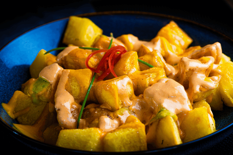A blue ceramic bowl filled with golden-yellow roasted potato cubes. They are drizzled with a creamy, pale orange sauce and garnished with thin red chili pepper slices and green chives under dramatic, low-key lighting.