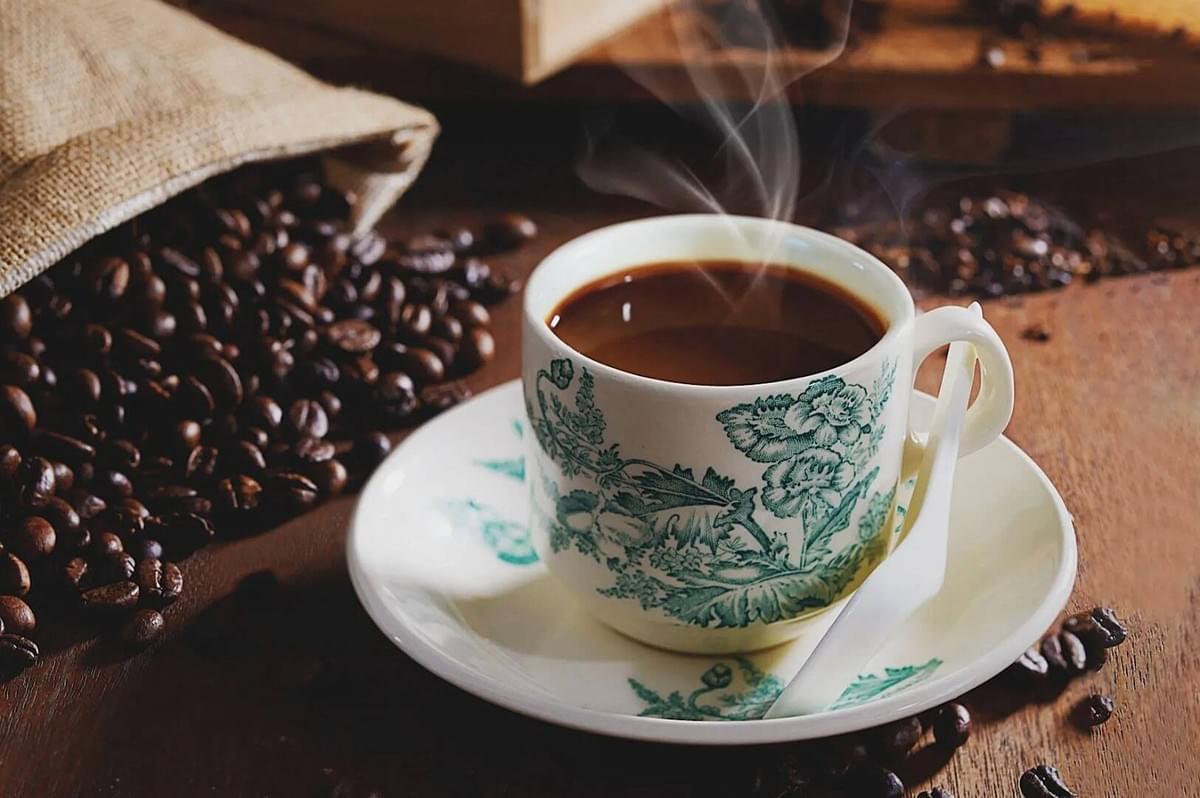 Steaming coffee in a floral-patterned cup on a saucer, surrounded by scattered coffee beans and a burlap sack, evoking a warm, cozy atmosphere.