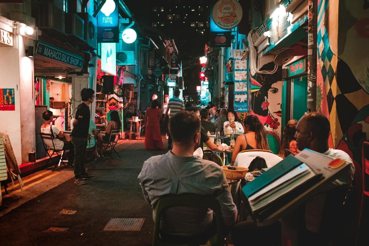 A bustling narrow alleyway comes alive at night with vibrant neon signs, street art, and people dining at outdoor tables. The warm glow of the shopfronts and string lights creates a lively, social atmosphere in this urban setting.