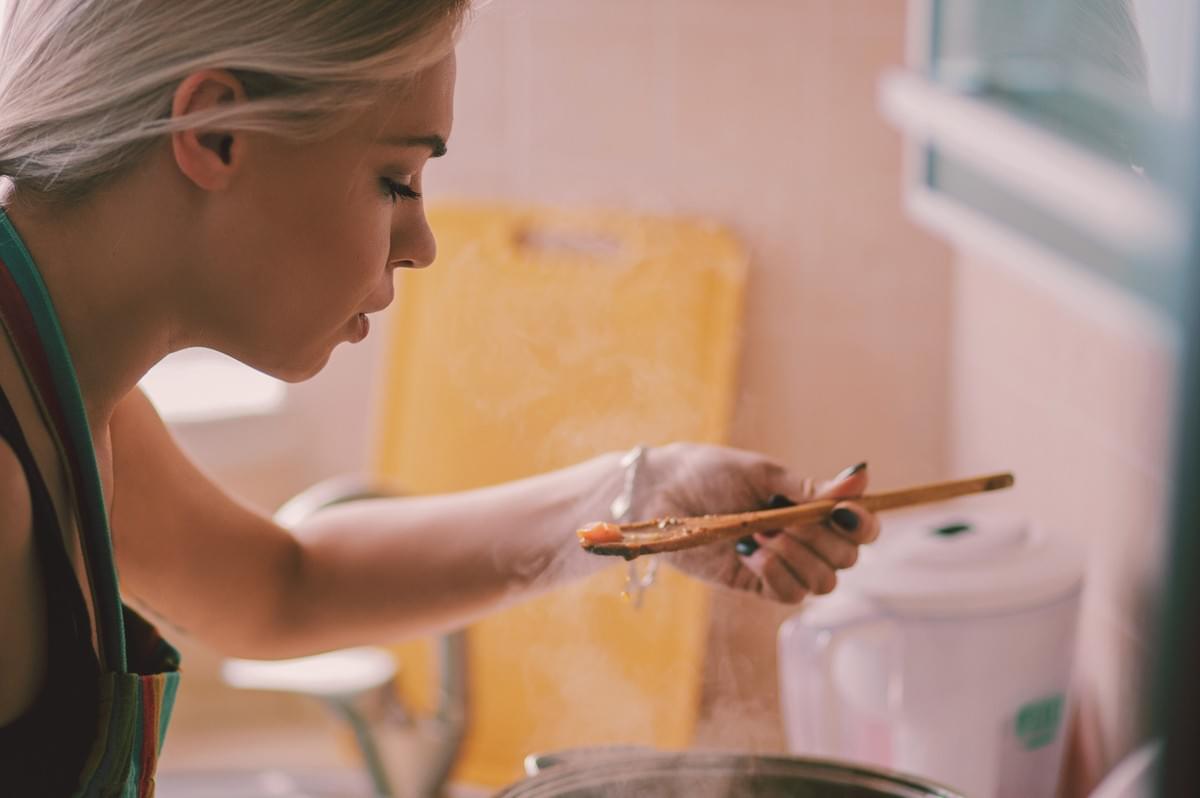 Person sampling a simmering dish with a wooden spoon over a steaming pot in a home kitchen.