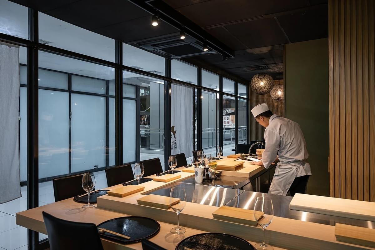 A sushi chef in white uniform prepares food behind a sleek, modern dining counter set with wine glasses and placemats. Large glass windows and warm pendant lights line the background of the elegant restaurant interior.