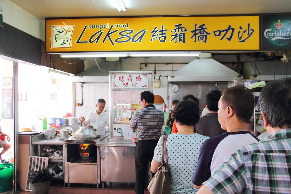 A long queue of customers waits in line at a Sungei Road Laksa food stall. Behind the counter, a cook prepares food over a traditional charcoal fire.