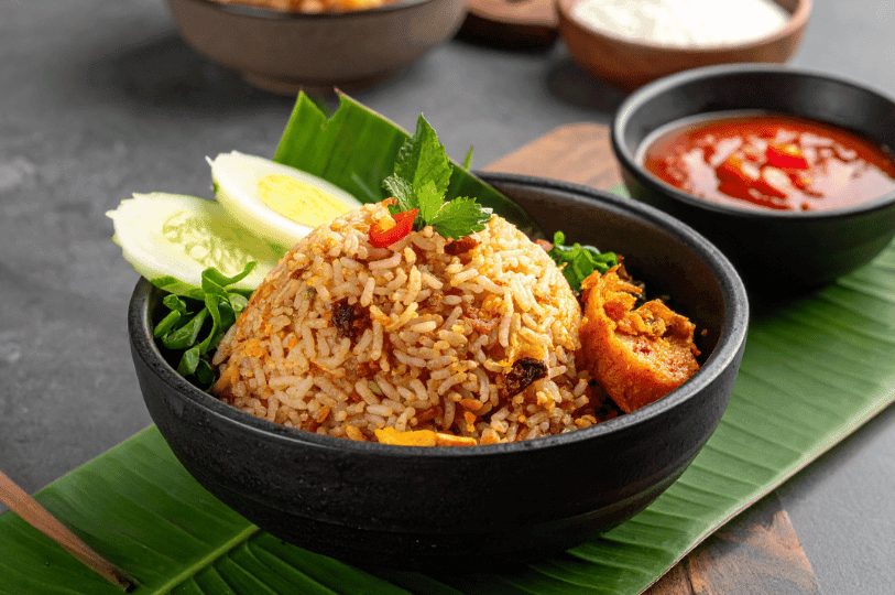 A rustic presentation of Nasi Goreng in a black ceramic bowl lined with a banana leaf, featuring aromatic rice, garnishes of fresh herbs, sliced cucumbers, and a side bowl of vibrant red chili sambal.