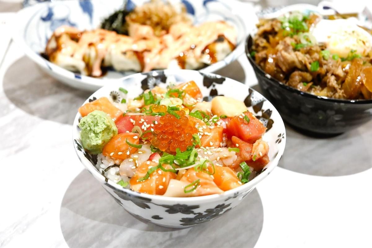 A vibrant bowl of bara chirashi don filled with cubed salmon, tuna, scallop, wasabi, and topped with salmon roe and chopped scallions sits in the foreground. In the background, a bowl of beef gyudon with a poached egg and another dish drizzled with sauce rest on a marble table.