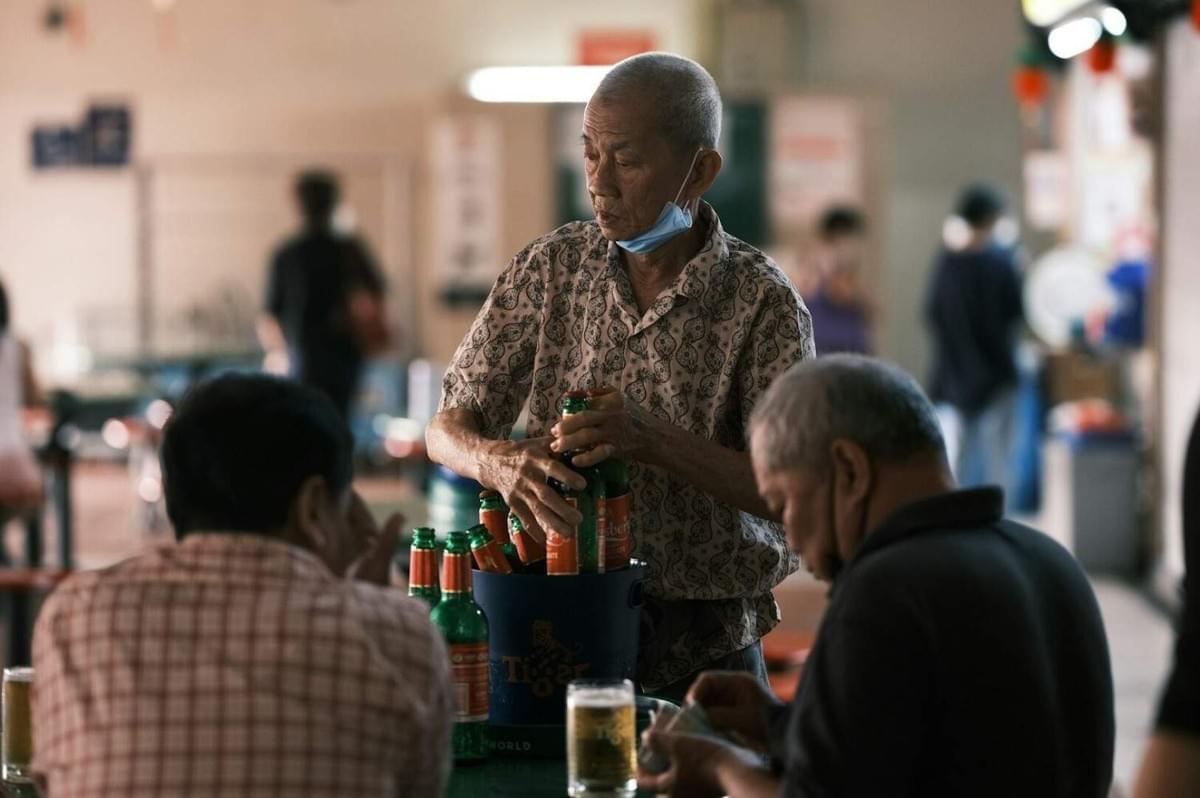 An elderly man wearing a face mask and holding a beer bottle, showcasing the essence of Singapore's hawker culture.