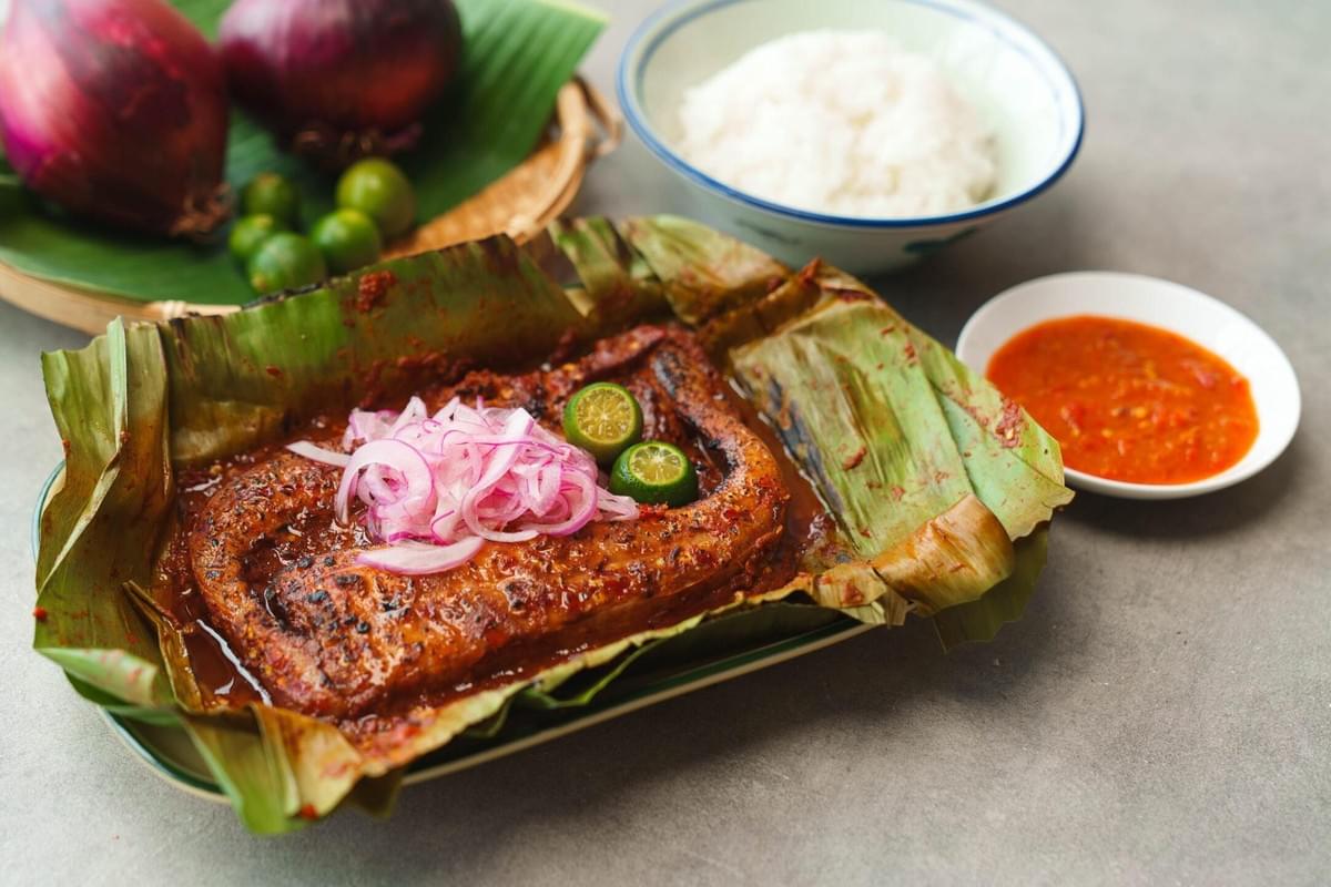 A grilled stingray wing coated in spicy sambal sits nestled in a charred banana leaf, topped with sliced red onions and calamansi lime. The meal is accompanied by a bowl of steamed white rice and a side of extra chili dipping sauce.