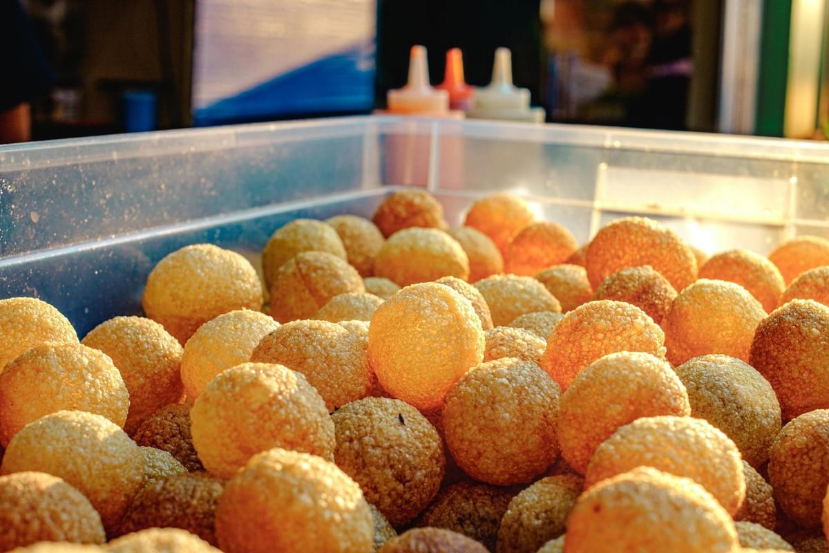 A clear plastic container is filled to the brim with numerous golden-brown, hollow, crispy puris used for making panipuri. In the blurred background, squeeze bottles containing various sauces or flavored waters stand ready for serving.