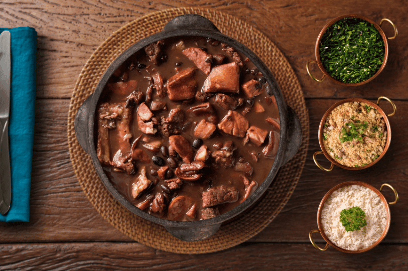 A traditional Brazilian feijoada served in a black pot, surrounded by copper bowls of rice, collard greens, and farofa—presented on a rustic table setting that evokes warmth, heritage, and communal celebration.