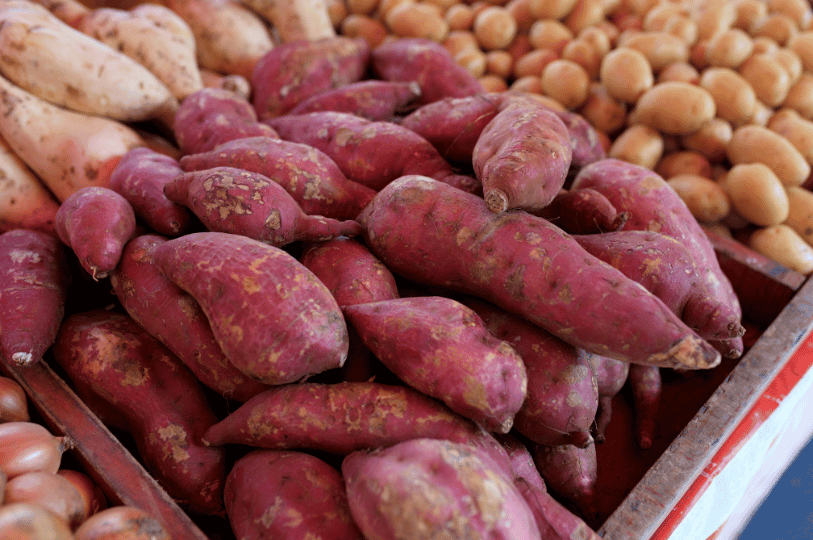 A high-angle shot of a market display featuring bins of reddish-purple sweet potatoes alongside regular white potatoes and onions.