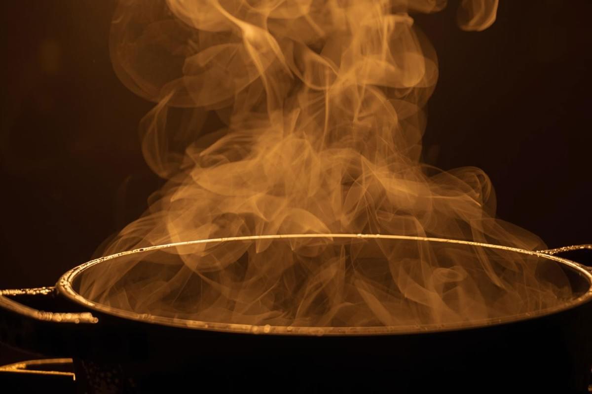 A dramatic close-up of a dark cooking pot with thick, golden-brown steam billowing up, suggesting high heat and a simmering liquid.