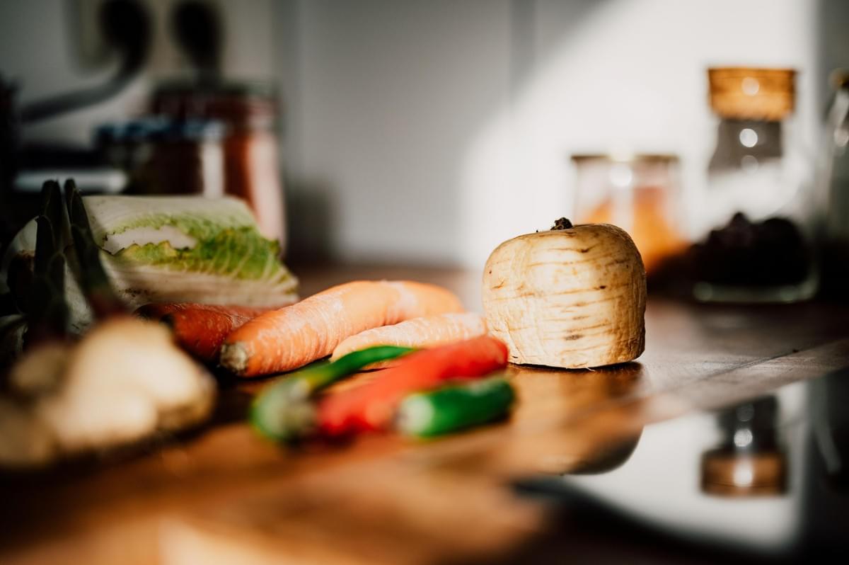 A collection of fresh vegetables, including carrots, leafy greens, a turnip, and chili peppers, sits arranged on a wooden kitchen countertop. The warm, natural light creates a soft focus on the blurred background filled with kitchen jars and utensils.