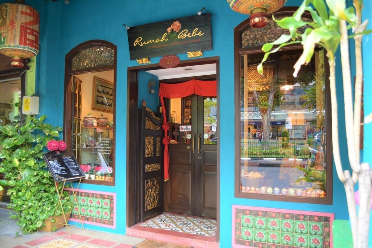 The entrance to Rumah Bebe is painted a vibrant turquoise and features ornate, gold-detailed wooden doors and traditional floral tiling along the base of the walls. Large display windows reveal heritage Peranakan items inside, while a decorative red fabric and a black menu board add to the shop's cultural charm.