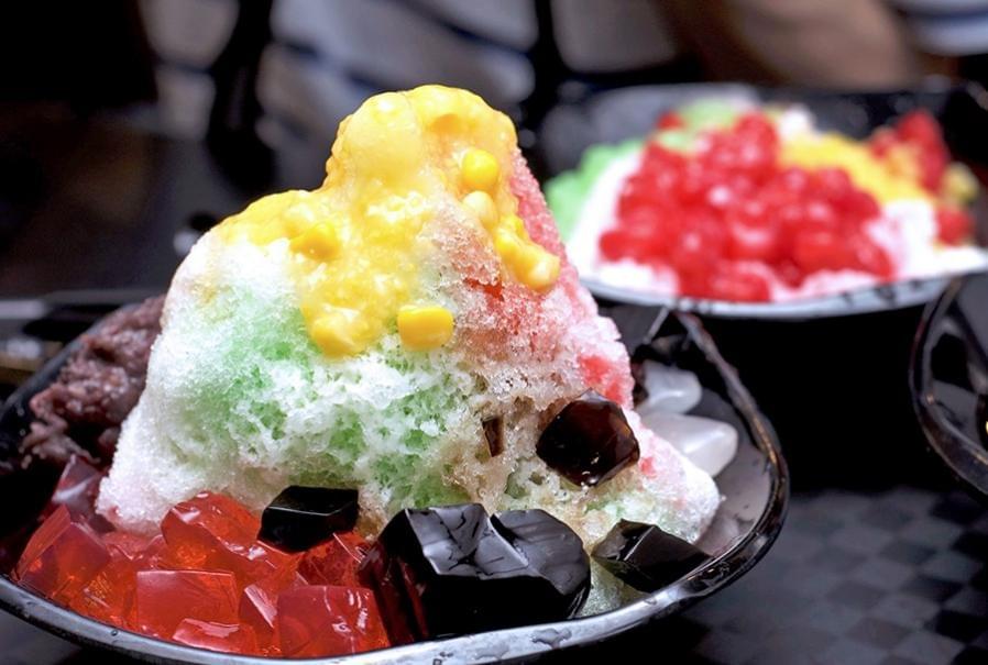 This image features a vibrant bowl of Ice Kacang, a popular shaved ice dessert topped with sweet corn, red beans, and colorful jellies. The refreshing treat is drizzled with bright syrups and served in a black bowl, highlighting the diverse textures of the ingredients.