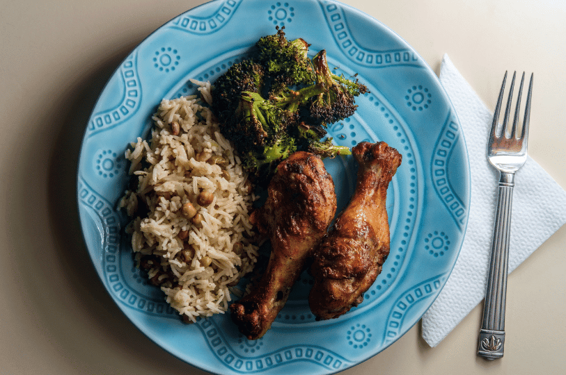 A vibrant blue plate featuring two grilled jerk chicken drumsticks, a side of traditional Jamaican rice and peas, and charred broccoli, served with a white napkin and silver fork.