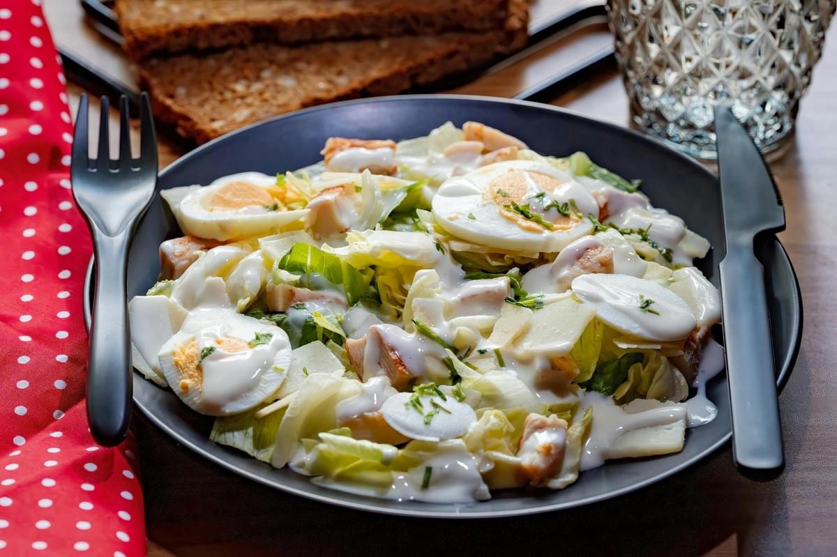 A dark plate holds a salad consisting of chopped lettuce, sliced hard-boiled eggs, chicken pieces, and cheese shavings, all topped with a white creamy dressing and fresh herbs. Black cutlery rests on the rim of the plate, which is set on a wooden surface beside a red polka-dot napkin and a textured glass.