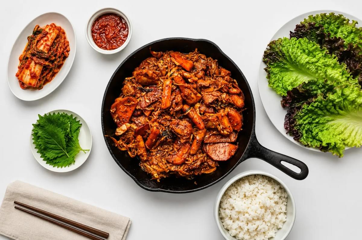 A flat-lay view of Korean dak-galbi (spicy stir-fried chicken) in a black cast-iron skillet, surrounded by side dishes of white rice, lettuce, perilla leaves, kimchi, and ssamjang paste on a white background.