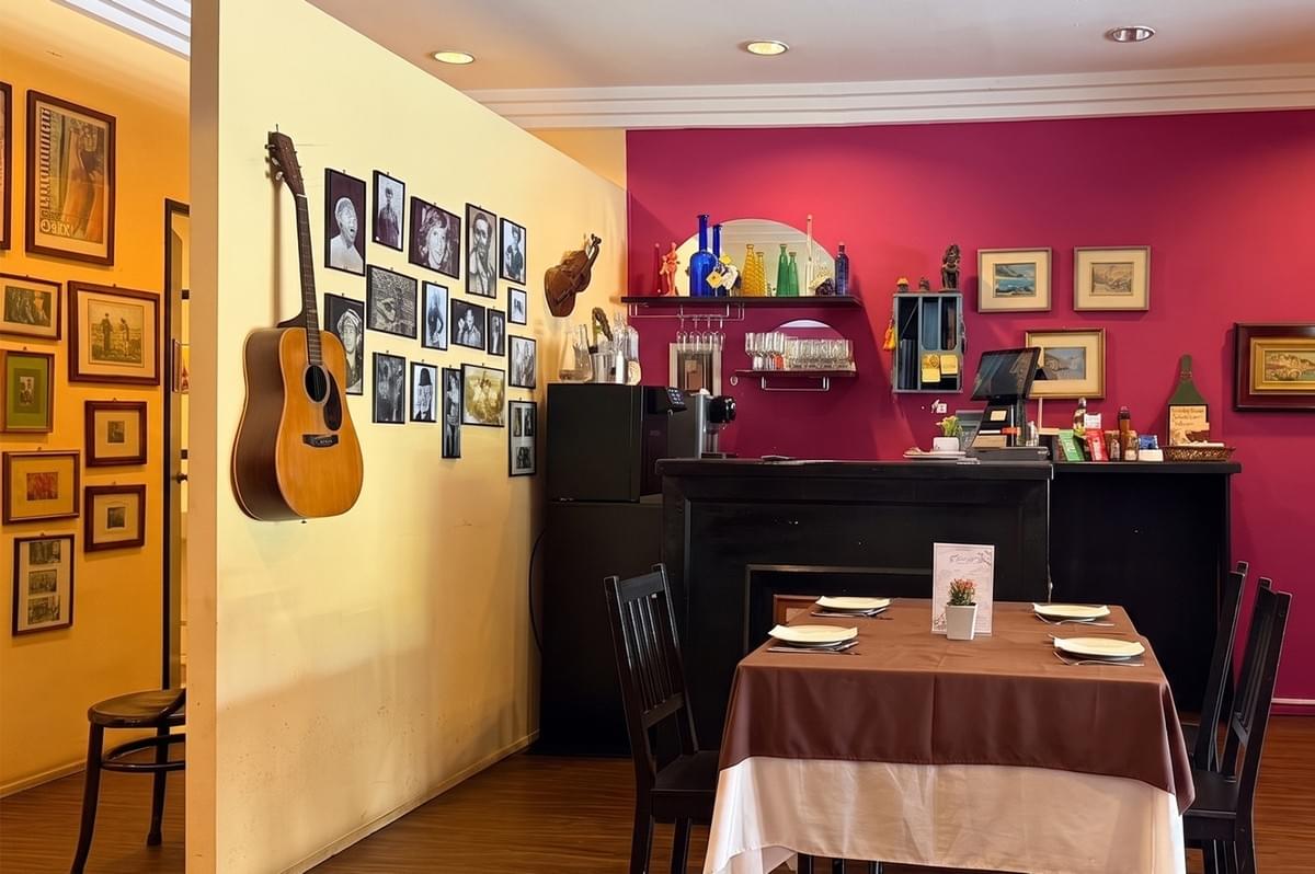 Interior view of a restaurant with a yellow wall featuring a guitar and framed photos, and a deep red wall with a service counter and dining table.