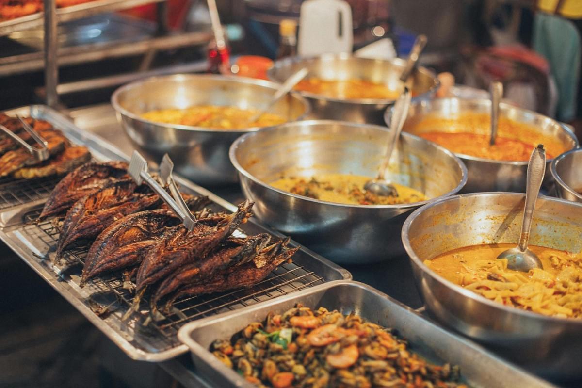 A vibrant street food stall displays various metal bowls filled with colorful, aromatic curries and stews. Alongside the bowls, trays of fried whole fish and stir-fried seafood are arranged for customers to choose from.