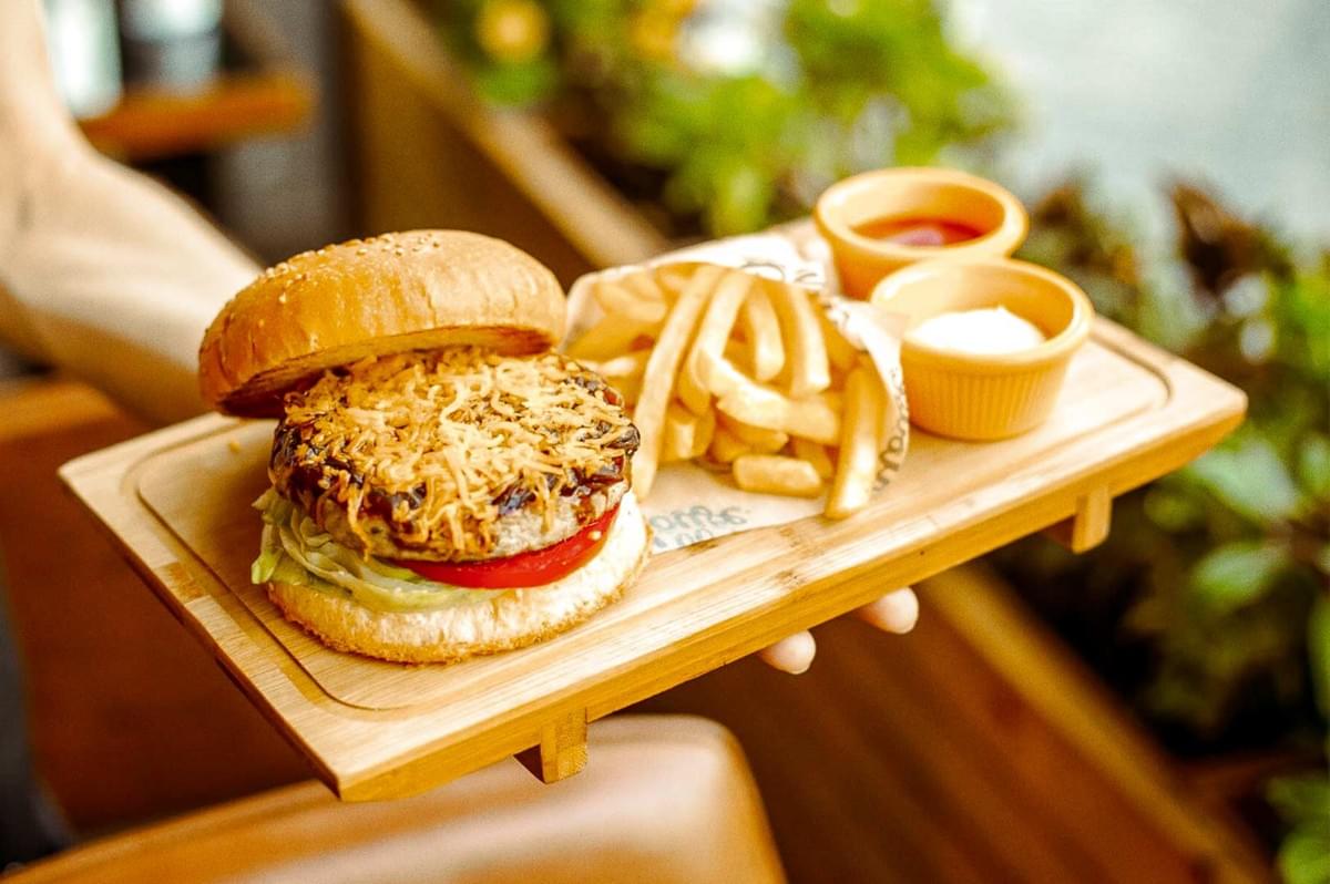 Cheeseburger on a sesame bun with shredded cheese, lettuce, and tomato served with paper-wrapped French fries and two dipping sauces on a wooden tray in a casual dining setting.