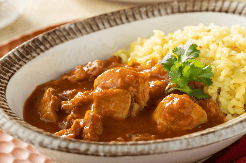 A ceramic bowl of butter chicken in rich reddish-brown sauce, served with vibrant yellow rice and cilantro garnish—celebrating the bold spices and warmth of Indian-inspired comfort food.