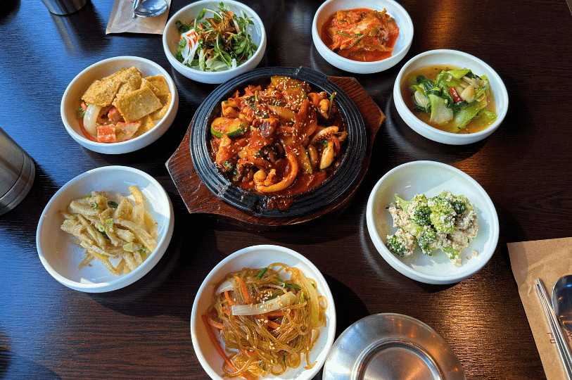A dark wooden table displaying a central hot iron skillet of spicy stir-fried squid, surrounded by six small bowls of complementary banchan, including slippery japchae glass noodles, stir-fried fish cakes, and a creamy broccoli salad.